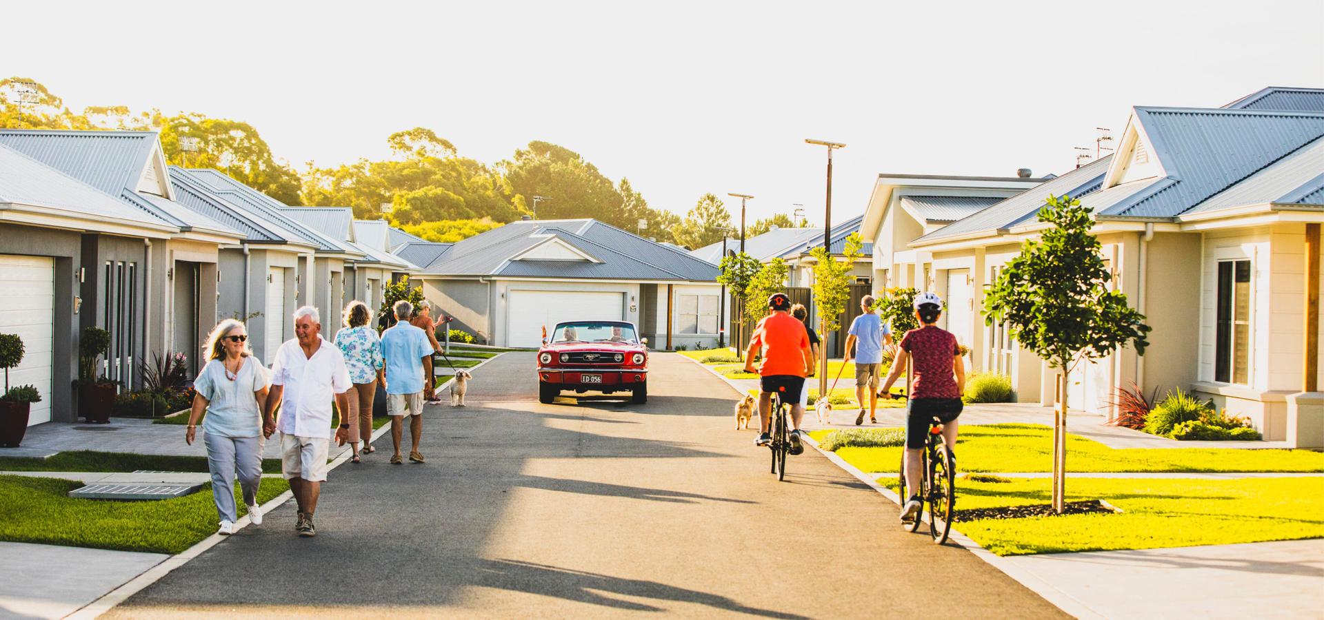 Sunlit suburban street in a lifestyle village featuring several houses, people walking/riding bikes (some with dogs), red convertible car driving by.
