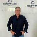 Man standing with hands on hips, wearing a dark shirt, in front of a white wall with logos/text possibly related to a lifestyle village amenity.