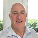 Smiling man with bald head in front of bright green foliage, wearing light blue collared shirt, with natural light creating a warm atmosphere.