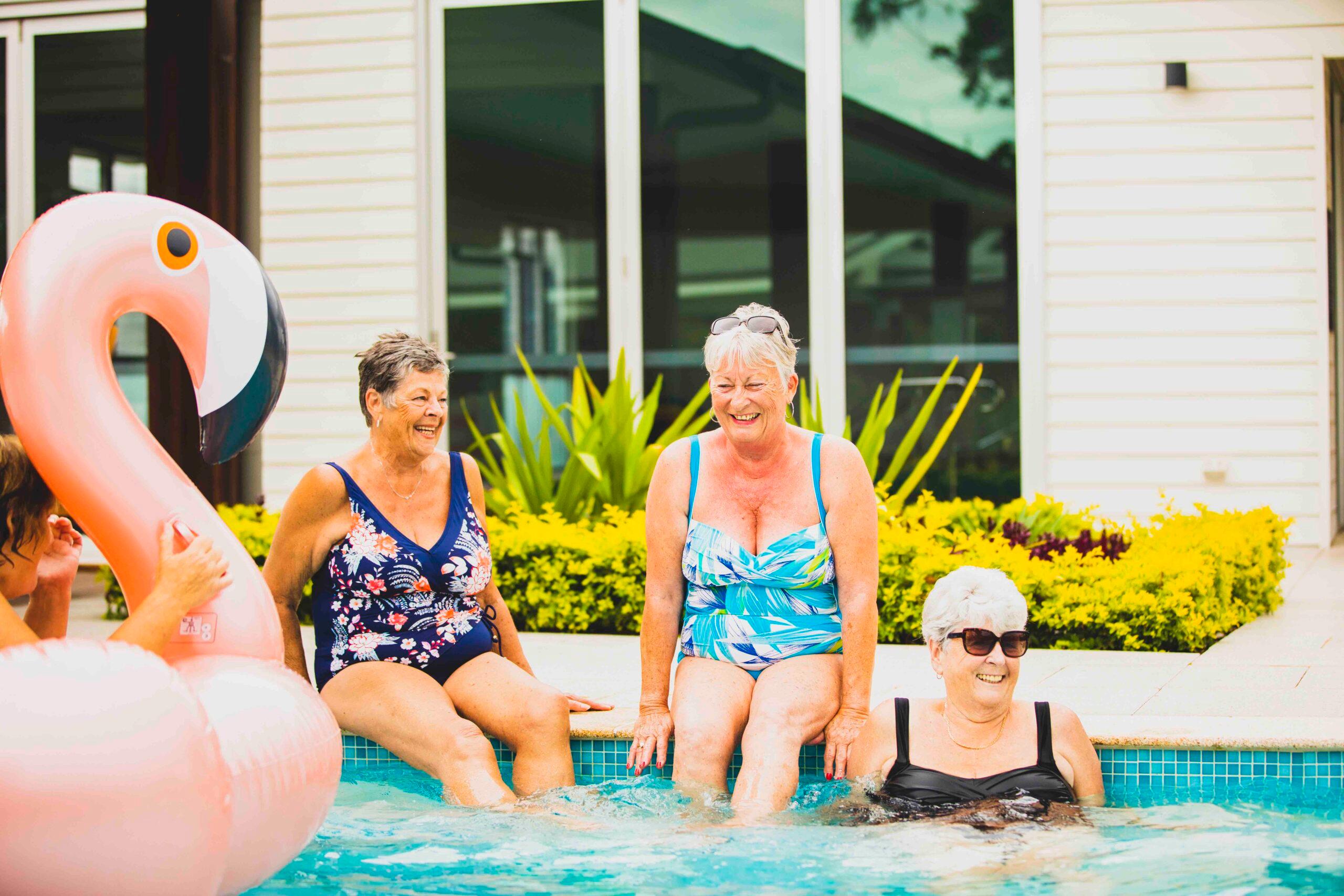 Four women in swimsuits sit edge of lifestyle village pool, playfully splashing water, large pink flamingo float partially visible, yellow/green plants surround.