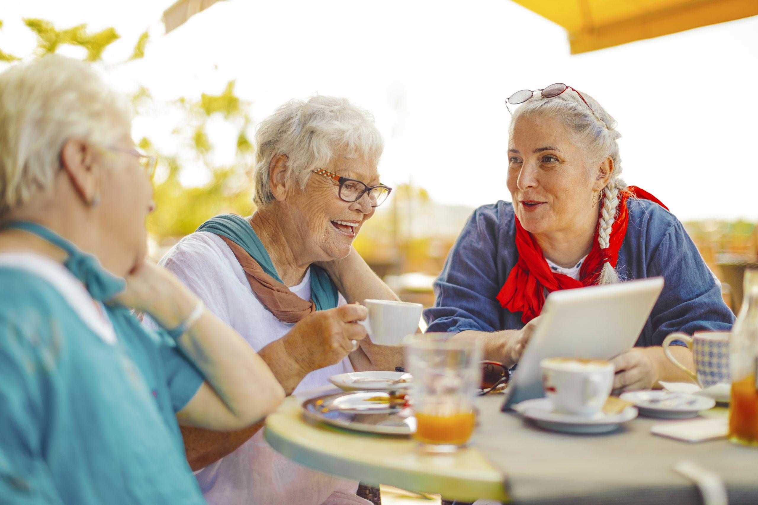 Three older women sit at lifestyle village café table, enjoying drinks/sharing laughter, one showing tablet, surrounded by food plates under sunny sky.