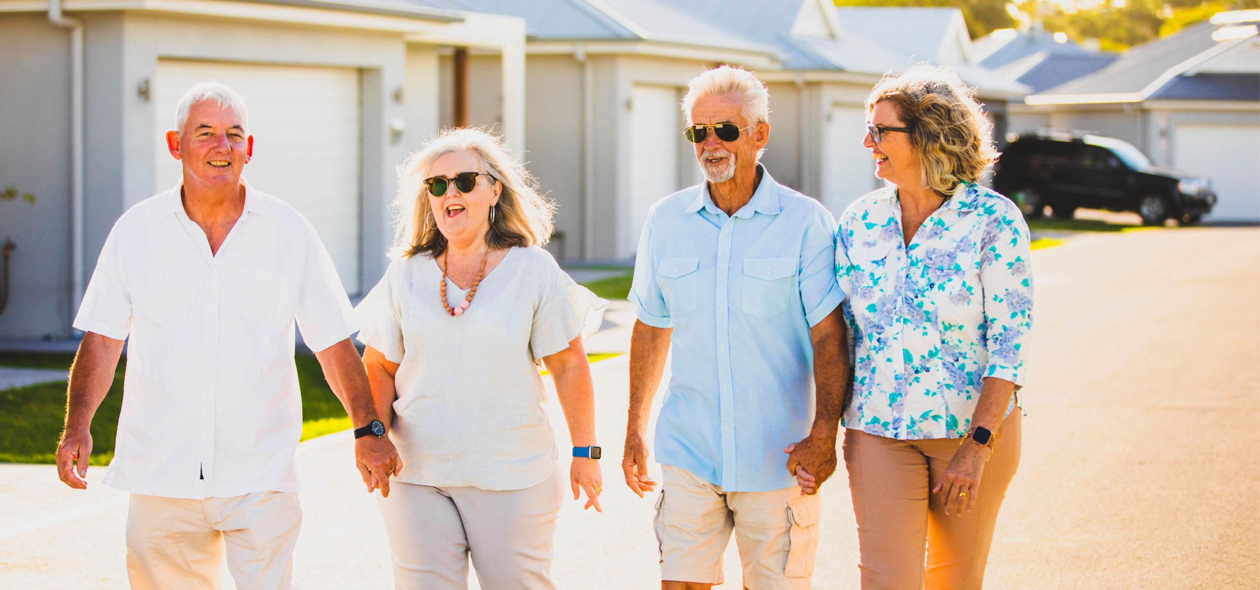 Four older adults walk together along sunny lifestyle village street, smiling enjoying each other’s company, wearing casual clothing/sunglasses, suburban homes.