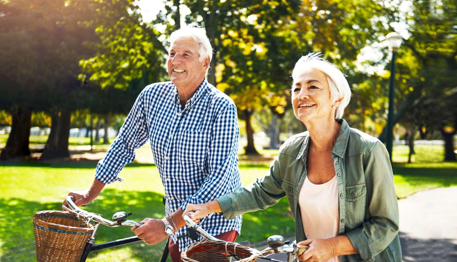 Two elderly people smiling walking beside their bicycles in lifestyle village park, surrounded by lush greenery/trees, reflecting warm sunny atmosphere.