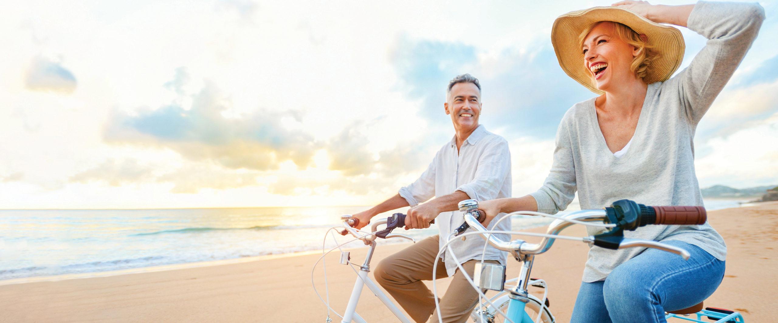 Two cyclists share a joyful moment on a beach, laughing while seated on their bicycles. The sun sets in the background, casting warm hues across the tranquil ocean.