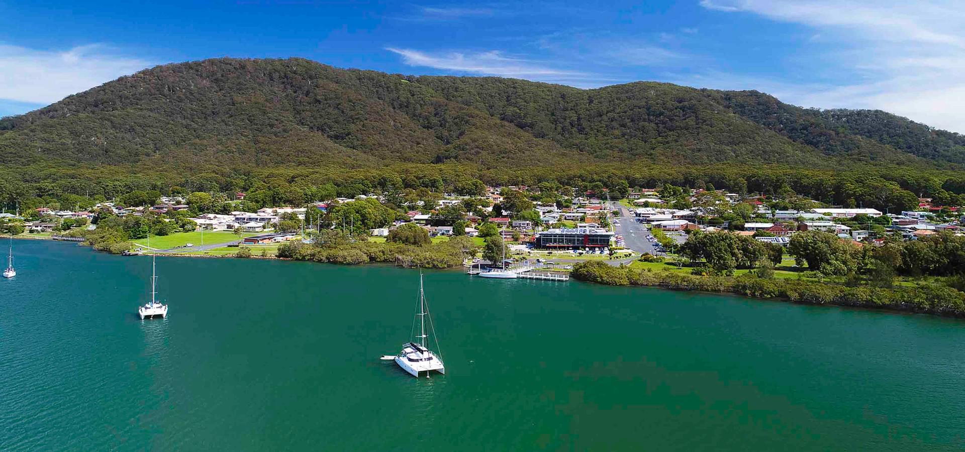 Sailboats float on calm green river, village sits at foot of lush forested mountain under clear blue sky, surrounded by greenery/buildings.