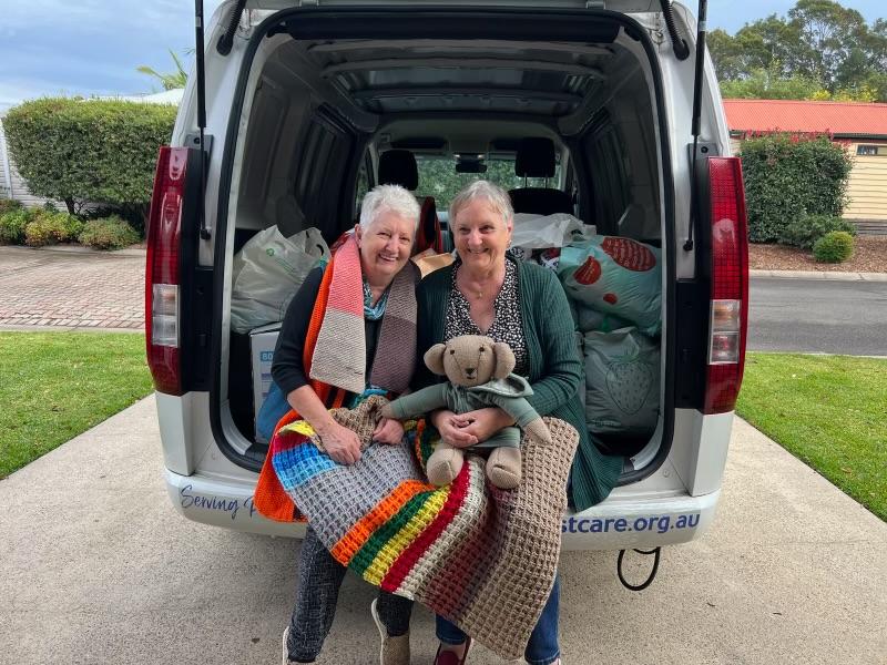 Two women sit together open back van, draped colorful blankets, hold stuffed toy, surrounded bags/boxes against grassy backdrop (possible community event).