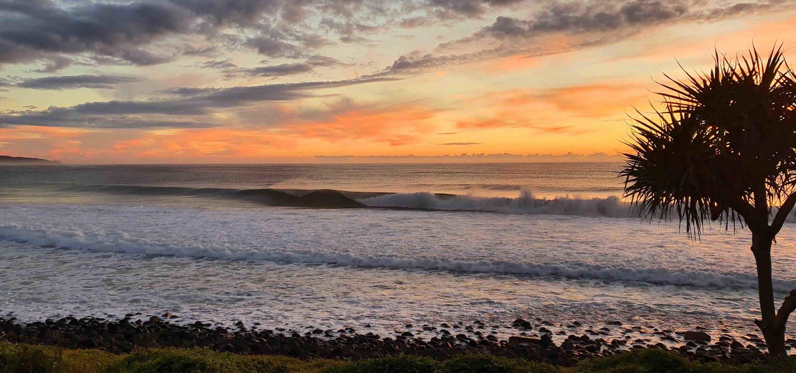 Waves roll onto sandy shore crashing gently against rocks, palm-like tree nearby silhouetted against vibrant sunset sky filled orange/pink clouds.