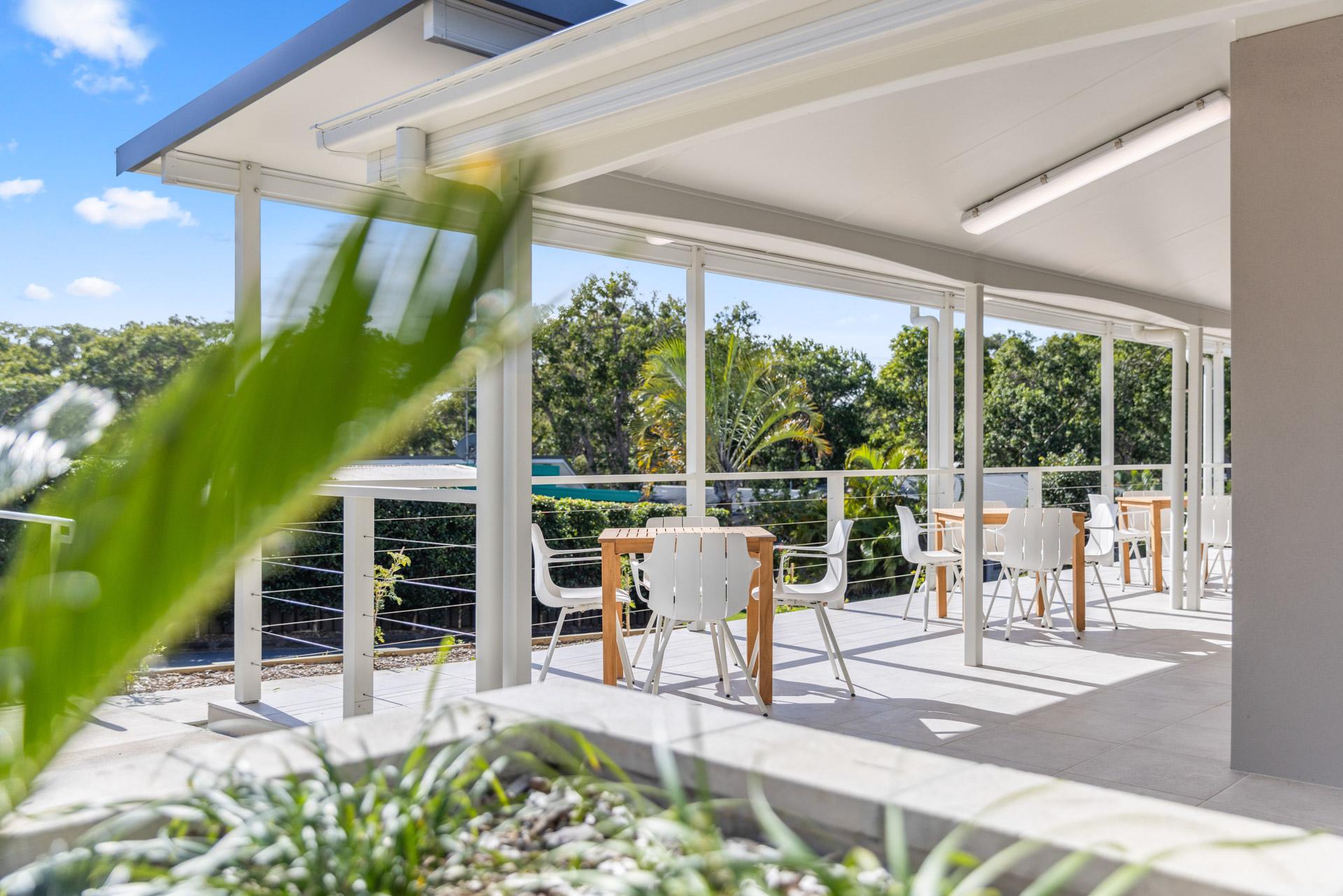 Relaxed outdoor dining patio in a lifestyle village featuring white tables and chairs, surrounded by lush greenery under bright daylight and clear blue skies.