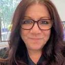 Woman (Tina) with long brown hair/glasses smiles at camera indoors, bright softly lit background with blurred greenery/colorful elements (lifestyle village context).
