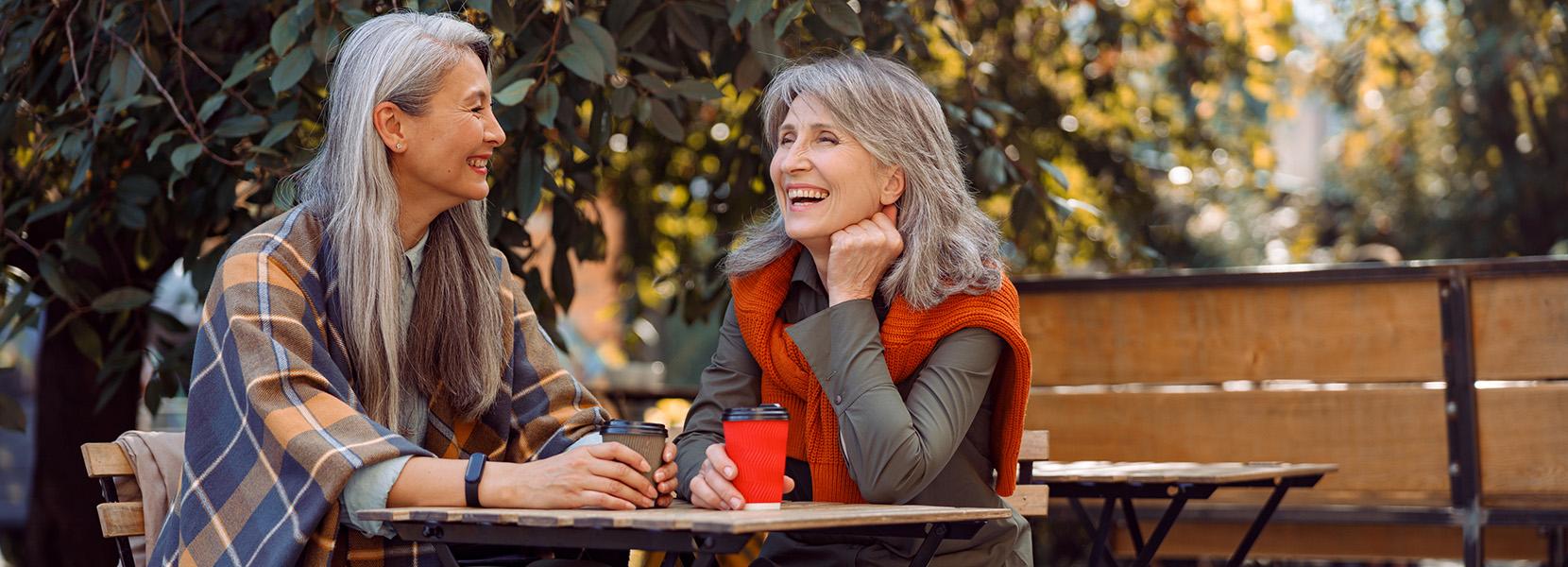 Two women with gray hair sit at outdoor table in lifestyle village park, smiling talking holding coffee cups, lush greenery surrounds, warm inviting atmosphere.