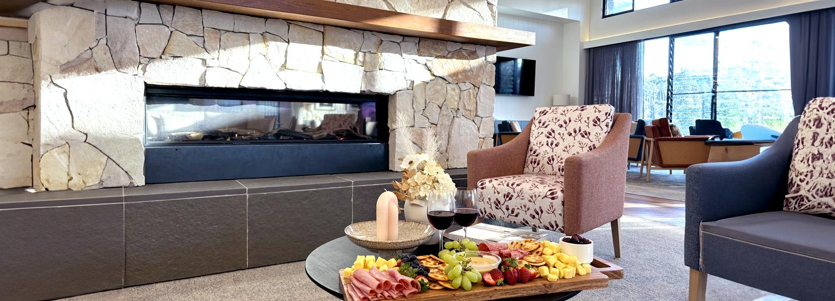 Wooden table displays platter fruits/cured meats, two glasses red wine nearby, modern fireplace warms cozy lifestyle village lounge area with plush chairs behind.