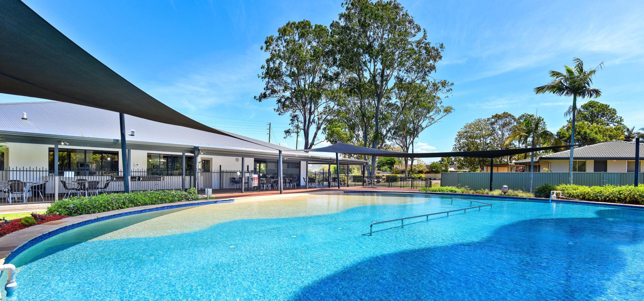 Clear lifestyle village swimming pool with a gradual shallow area, near a building with large windows, shade sails over lounge chairs, and lush greenery.