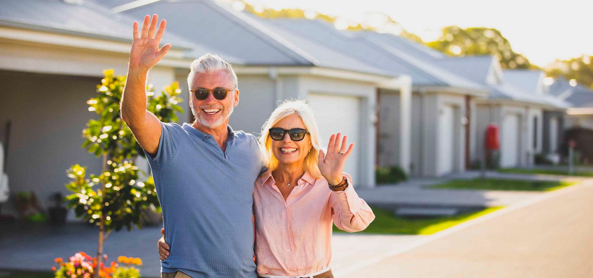 Smiling couple waving joyfully while standing outdoors in front of modern lifestyle village homes, wearing sunglasses under a clear blue sky.