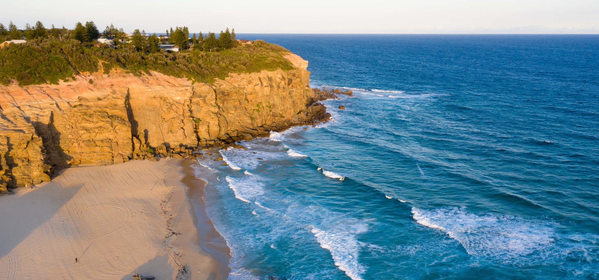 Cliffs rise over sandy beach where gentle waves ebb/flow, people strolling shore, lush greenery/distant homes line cliff edge under clear blue sky.