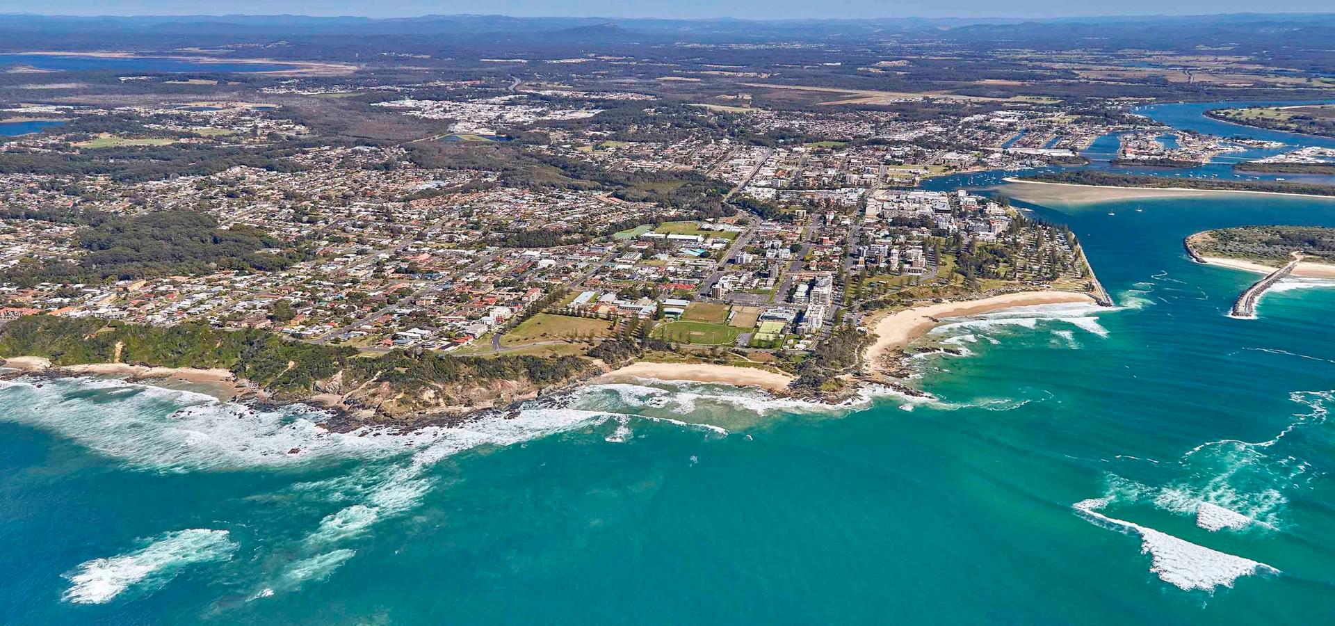 Aerial view of a coastal town showing residential areas, green spaces, sandy beaches with crashing waves, hills, and distant fields.
