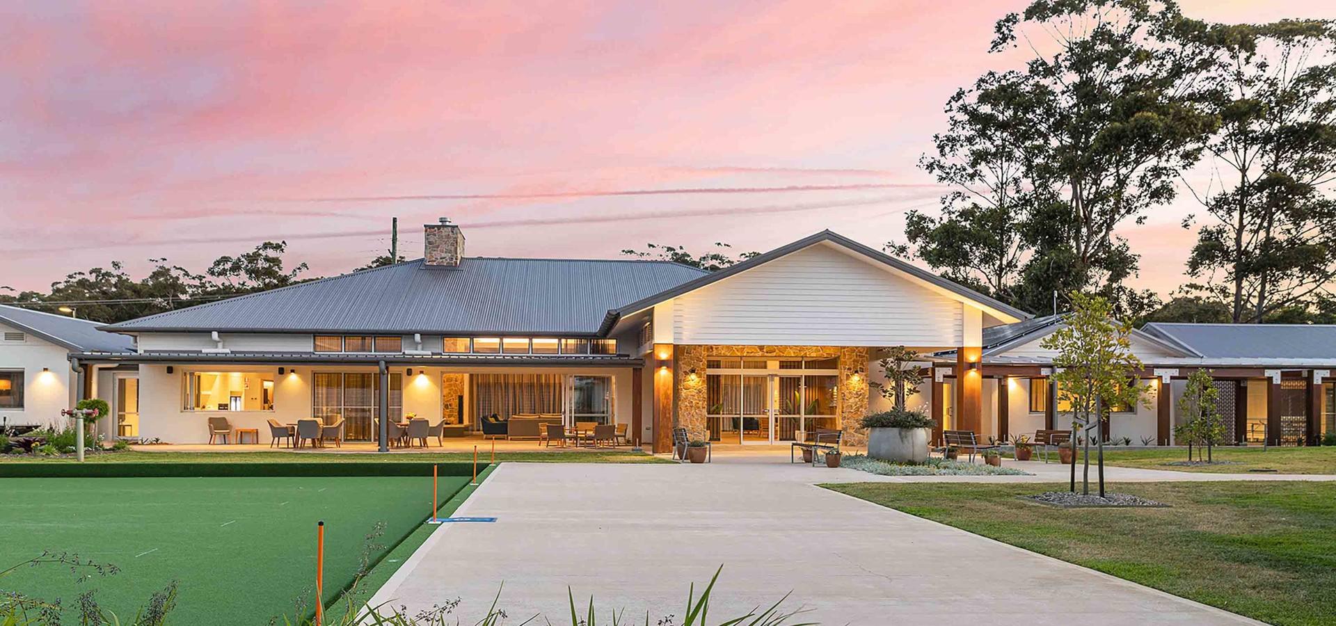 Modern, single-story lifestyle village building with large windows and stone entrance, amidst green grass and trees under a colorful twilight sky.