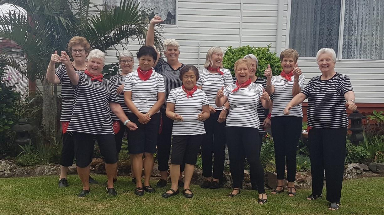Group of nine women in matching striped shirts and black pants smiling with thumbs up in a lifestyle village yard surrounded by greenery.